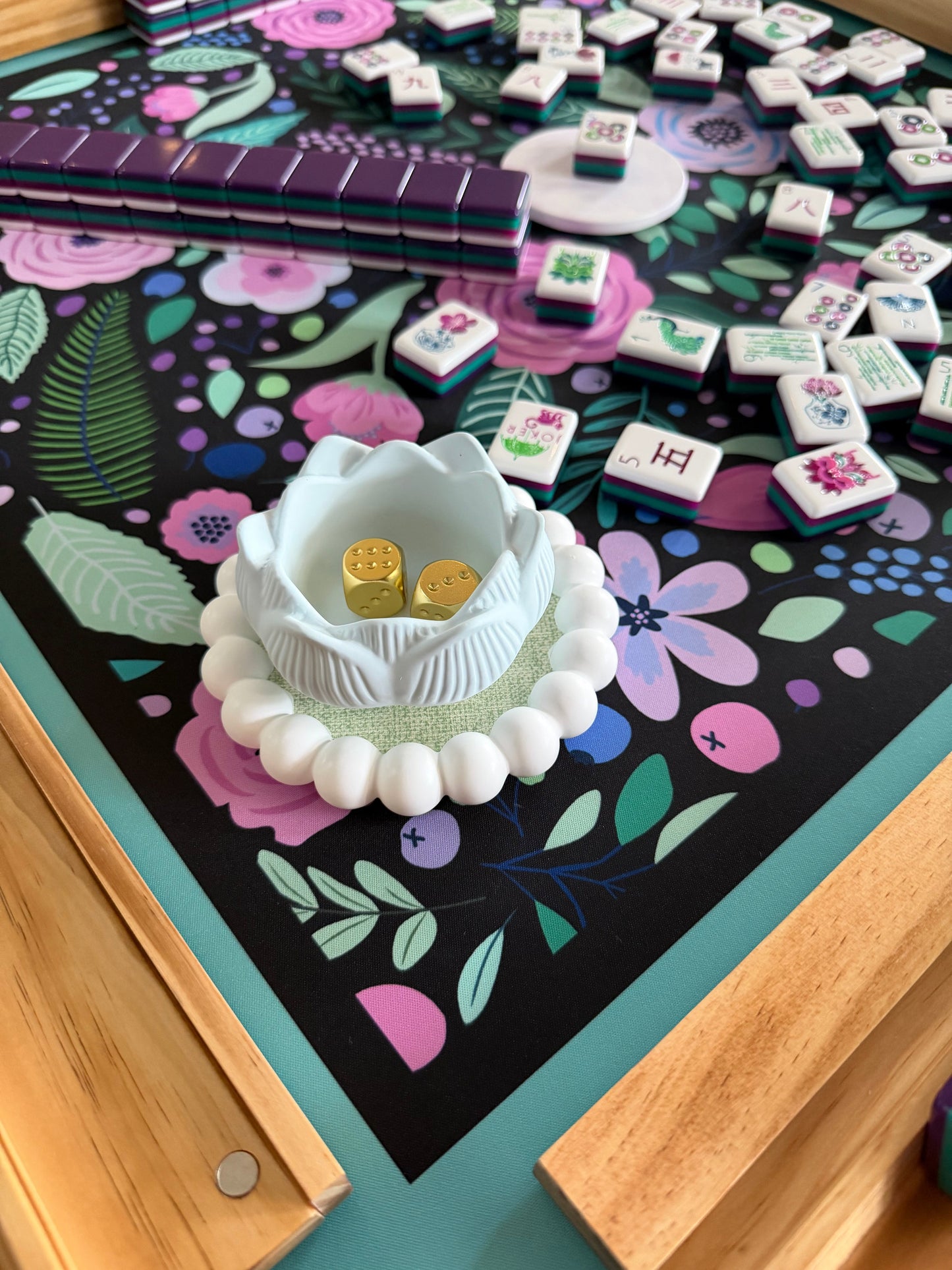 A white lotus dish holds gold dice on a floral mahjong mat scattered with tiles and a wood rack