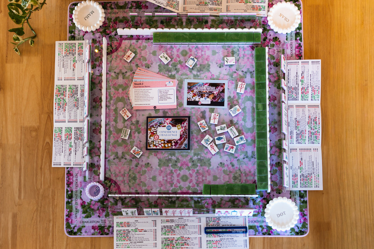 Overhead mahjong table setup with a floral mat, tile racks, course cards, and a video preview screen.